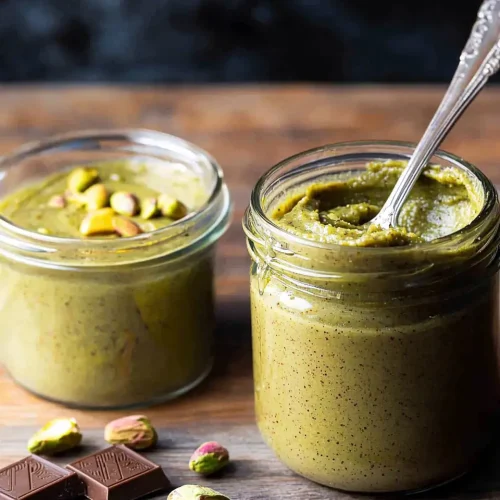 Best Pistachio Butter for Dubai Chocolate 5 best pistachio butter for dubai chocolate Beautiful jar of homemade pistachio butter with fresh pistachios and Dubai chocolate pieces on rustic wooden background