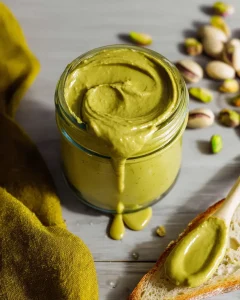 Glass jar of pistachio butter recipe for dubai chocolate with wooden spoon and spread on bread showing creamy green texture