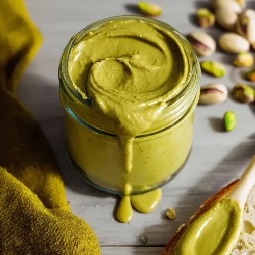 Glass jar of pistachio butter recipe for dubai chocolate with wooden spoon and spread on bread showing creamy green texture