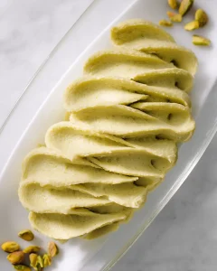 Bowl of smooth, pale green pistachio cream butter with offset spatula, surrounded by whole pistachios on marble surface