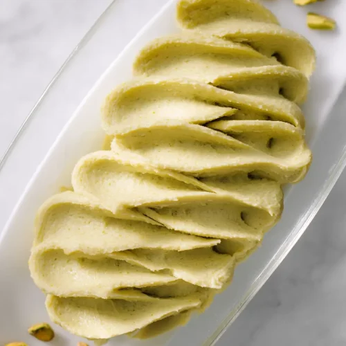 Bowl of smooth, pale green pistachio cream butter with offset spatula, surrounded by whole pistachios on marble surface
