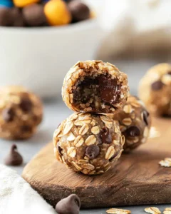 Stack of cookie balls no bake on wooden board with one cut in half showing oats, chocolate chips and M&Ms inside