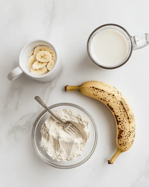 Three ingredients for protein powder mug cake arranged on white marble counter - protein powder in bowl, mashed banana, and glass of milk
