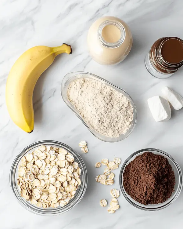 Ingredients for no bake protein cookies arranged on white marble counter including oats, protein powder, peanut butter, almond milk, banana, and cocoa powder